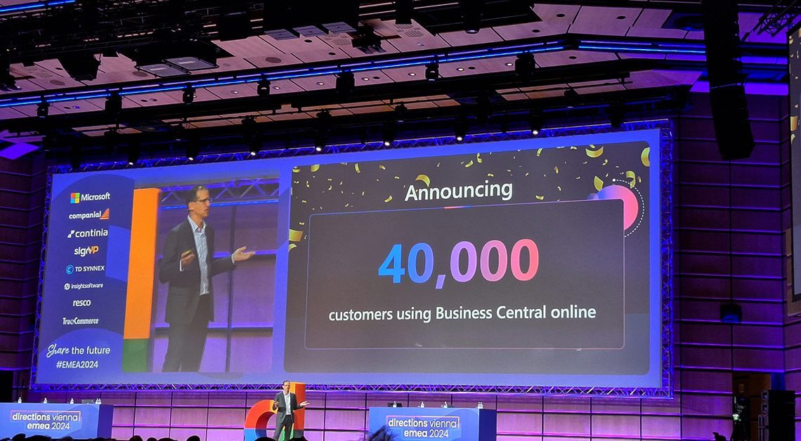 En stor skärm på Directions EMEA 2024 visar "Announcing 40,000 customers using Business Central online", och bredvid den står en presentatör och talar på scenen. Lokalen är väl upplyst och modern, med synliga företagslogotyper på skärmen.