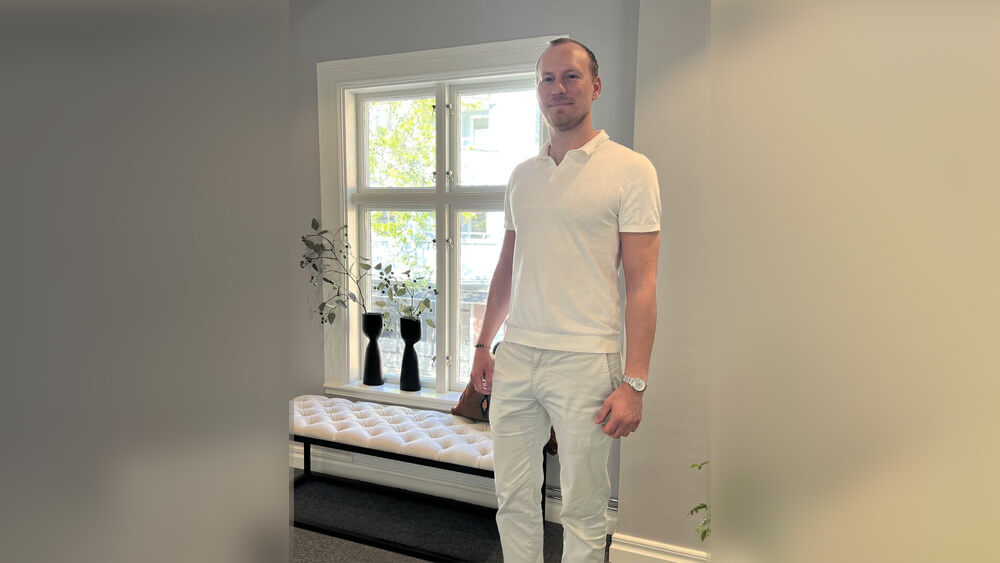 Välkommen Pål! En man i vit skjorta och byxor står inomhus vid ett fönster med vita karmar. Bakom honom står en vit kuddbänk och två svarta vaser med grenar, allt i naturligt ljus från det mjuka, ljusa rummet.