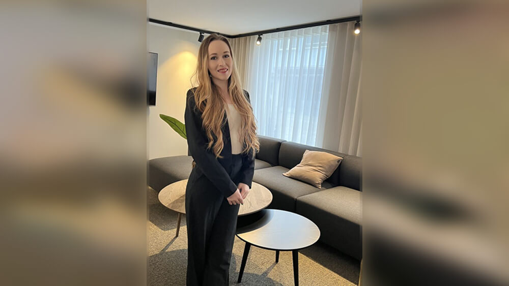 Välkommen Hanna - en kvinna med långt, vågigt blont hår i svart kostym står leende i ett modernt kontor med mörk soffa, runda bord, ljusa gardiner och dämpad belysning.