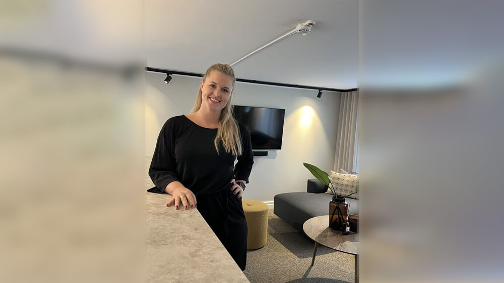 Välkommen Jessica! En kvinna med långt blont hår, klädd i en svart outfit, står och ler i ett modernt vardagsrum med en väggmonterad TV, grå soffa, gul ottoman och prydnadsföremål på ett bord.