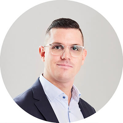En man med kort mörkt hår, som bär klara glasögon, en vit skjorta med krage och en mörk kavaj, poserar mot en ljusgrå bakgrund som representerar Yellow Solution när han tittar in i kameran med ett neutralt uttryck.