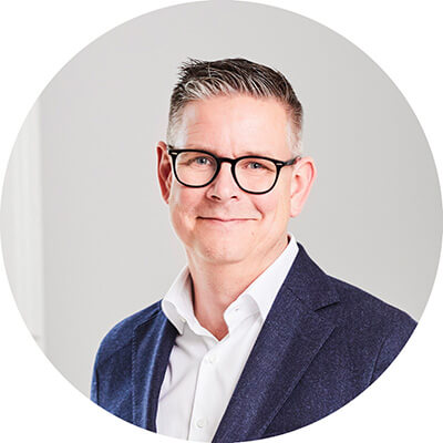 En man med kort grått hår och glasögon, klädd i en vit skjorta och en marinblå kavaj, ler mot kameran mot en ljus bakgrund och utstrålar självförtroende som någon som presenterar en gul lösning.
