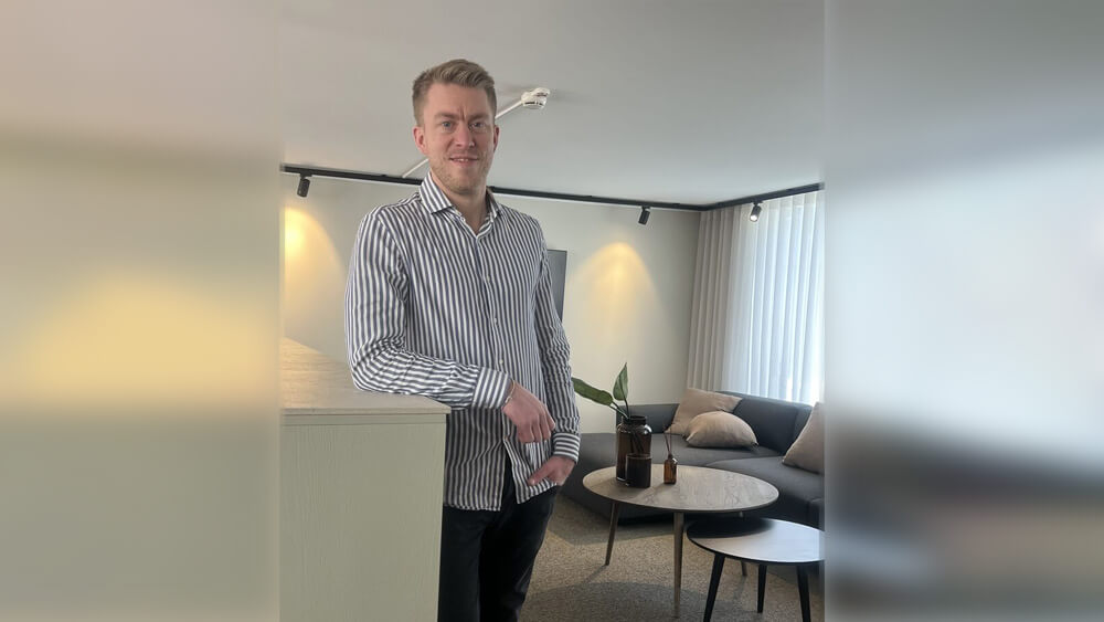 Välkommen Pål! En man i randig skjorta står och ler i ett modernt vardagsrum med soffa, kuddar, ett runt soffbord och krukväxter. Rummet är mjukt upplyst av naturligt ljus från höga fönster.