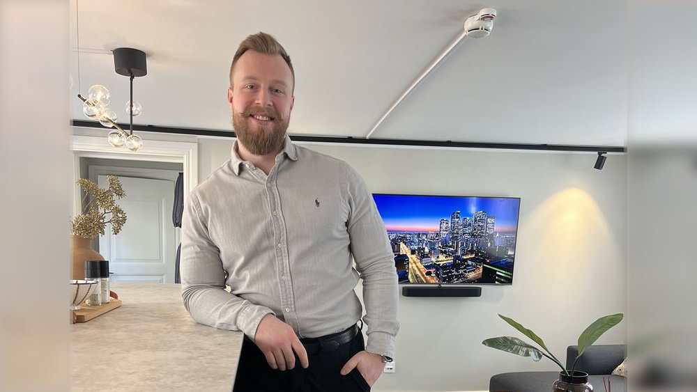 Välkommen Pål: En man med skägg, klädd i ljusgrå skjorta och svarta byxor, står och lutar sig mot en köksbänk i ett modernt vardagsrum. En TV på väggen visar en stadsbild i skymningen, medan växter och inredning ger rummet värme.