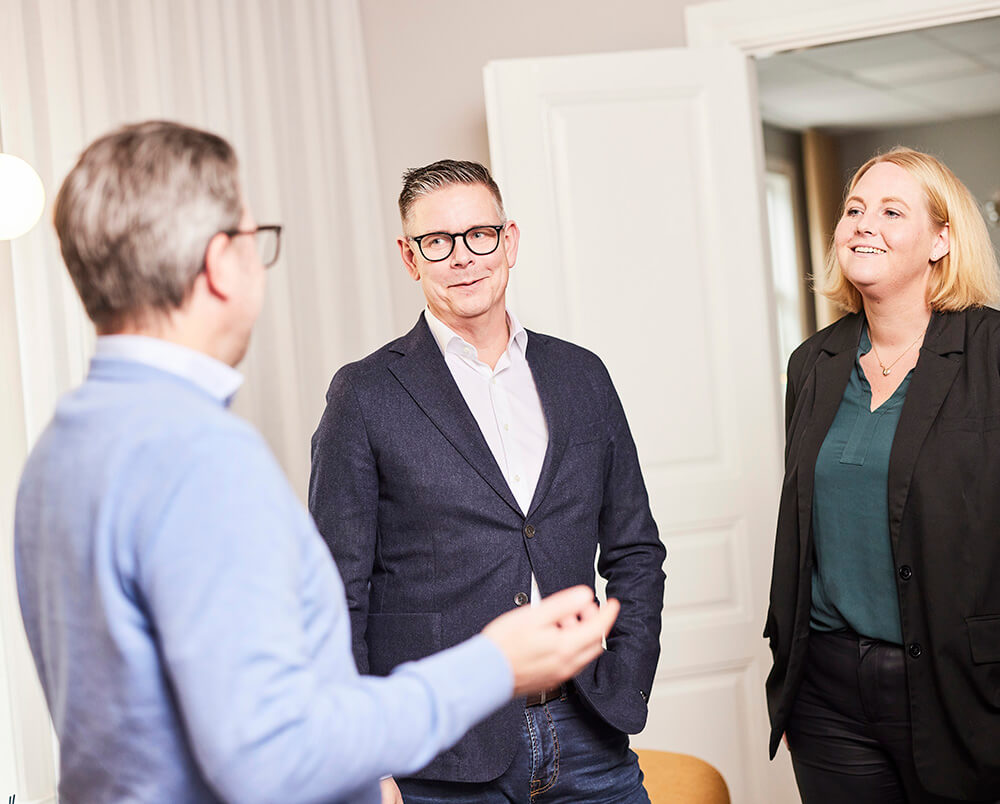 Tre personer i affärskläder står tillsammans inomhus och samtalar. Två män möter en kvinna, alla ser vänliga och professionella ut när de diskuterar en gul lösning. I bakgrunden syns vita väggar och en öppen dörr.