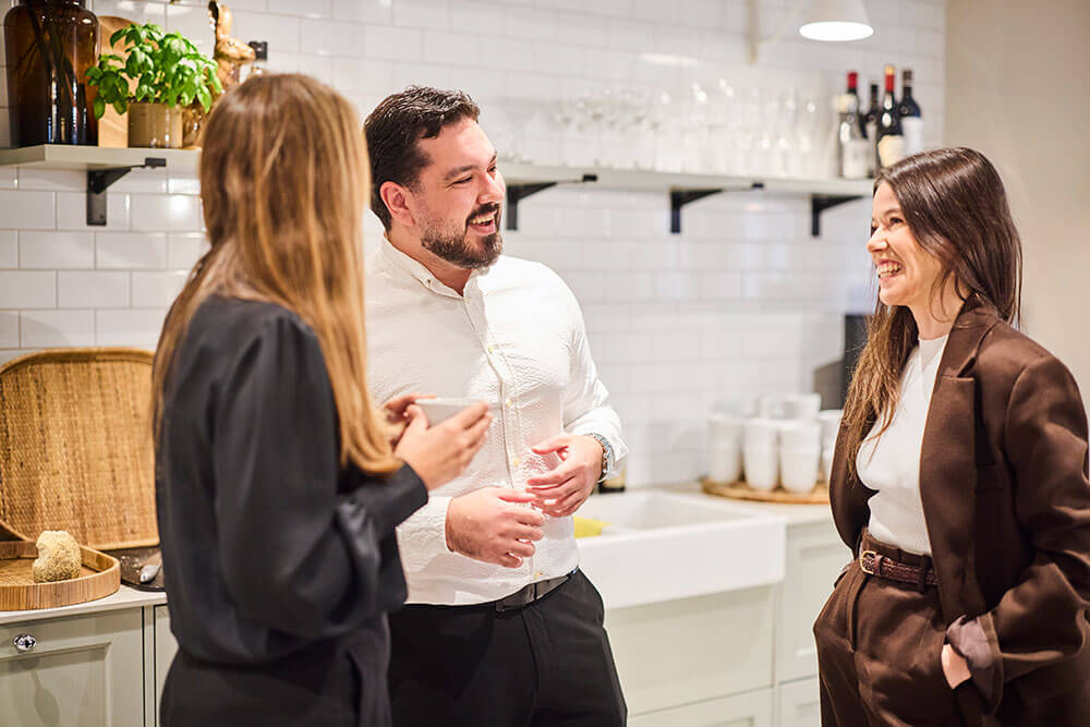 Tre personer står i ett ljust, modernt kök och ler och småpratar. Två kvinnor och en man ser ut att vara avslappnade och engagerade i en konversation med koppar i handen. Hyllor med glas och flaskor syns i bakgrunden.