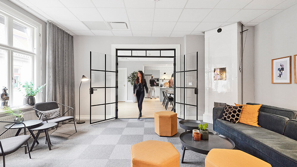 En modern kontorslounge med gråa soffor, gula ottomaner och runda bord välkomnar Utvecklare Business Centrals medarbetare. Stora fönster, växter och inramade konstverk skapar ett inbjudande utrymme när en kvinna går in genom dubbeldörrar i glas.