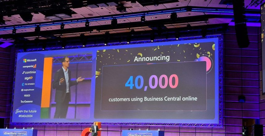 En stor skärm på Directions EMEA 2024 visar "Announcing 40,000 customers using Business Central online", och bredvid den står en presentatör och talar på scenen. Lokalen är väl upplyst och modern, med synliga företagslogotyper på skärmen.