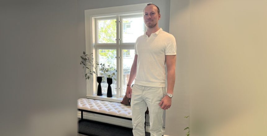 Välkommen Pål! En man i vit skjorta och byxor står inomhus vid ett fönster med vita karmar. Bakom honom står en vit kuddbänk och två svarta vaser med grenar, allt i naturligt ljus från det mjuka, ljusa rummet.