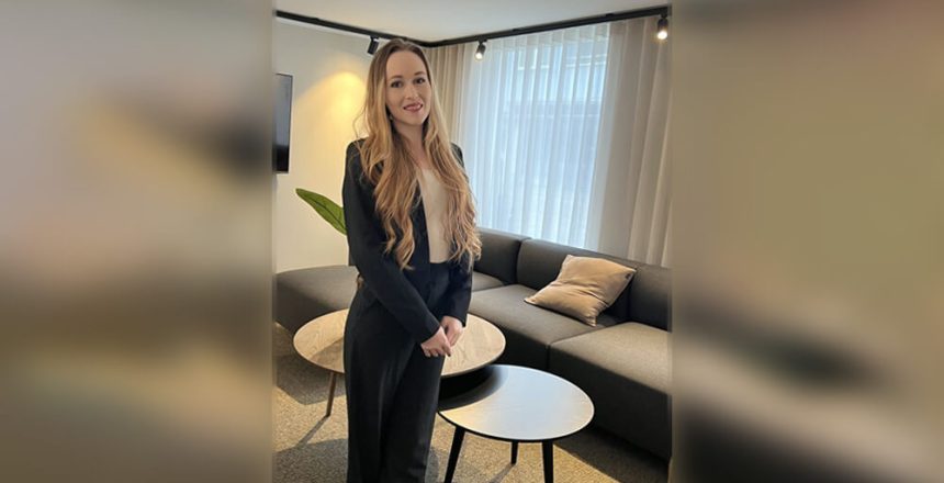 Välkommen Hanna - en kvinna med långt, vågigt blont hår i svart kostym står leende i ett modernt kontor med mörk soffa, runda bord, ljusa gardiner och dämpad belysning.