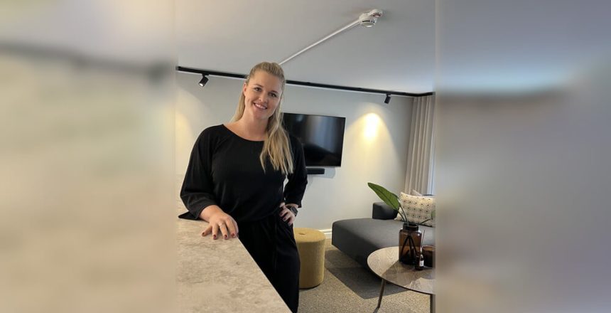 Välkommen Jessica! En kvinna med långt blont hår, klädd i en svart outfit, står och ler i ett modernt vardagsrum med en väggmonterad TV, grå soffa, gul ottoman och prydnadsföremål på ett bord.