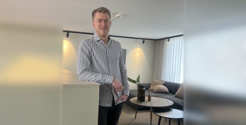 Välkommen Pål! En man i randig skjorta står och ler i ett modernt vardagsrum med soffa, kuddar, ett runt soffbord och krukväxter. Rummet är mjukt upplyst av naturligt ljus från höga fönster.