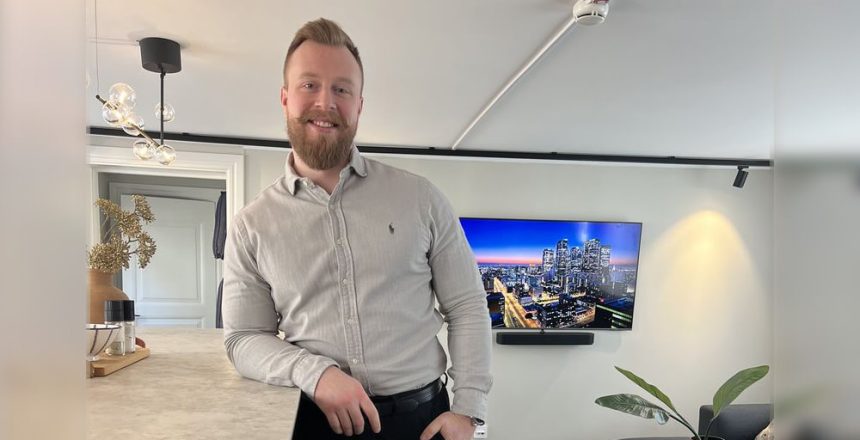 Välkommen Pål: En man med skägg, klädd i ljusgrå skjorta och svarta byxor, står och lutar sig mot en köksbänk i ett modernt vardagsrum. En TV på väggen visar en stadsbild i skymningen, medan växter och inredning ger rummet värme.