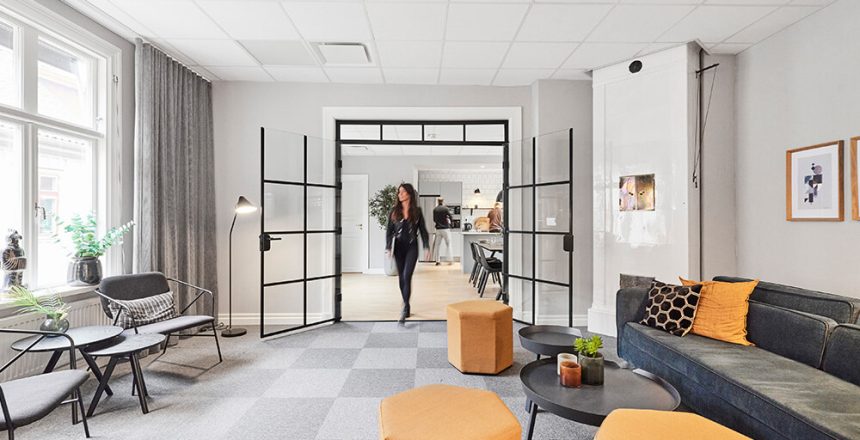 En modern kontorslounge med gråa soffor, gula ottomaner och runda bord välkomnar Utvecklare Business Centrals medarbetare. Stora fönster, växter och inramade konstverk skapar ett inbjudande utrymme när en kvinna går in genom dubbeldörrar i glas.