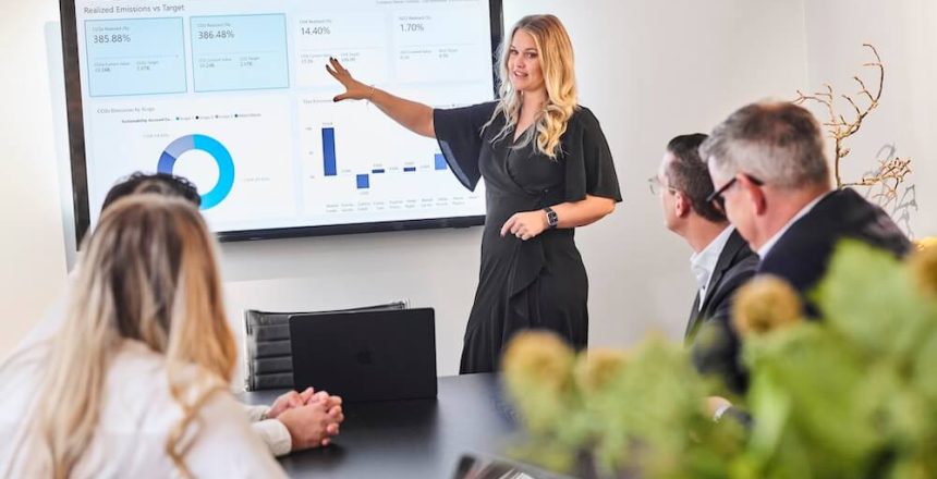 En kvinna står och pekar på en skärm som visar diagram och grafer under ett affärsmöte, medan tre personer som sitter vid ett bord lyssnar uppmärksamt. En bärbar dator och några växter syns i förgrunden.